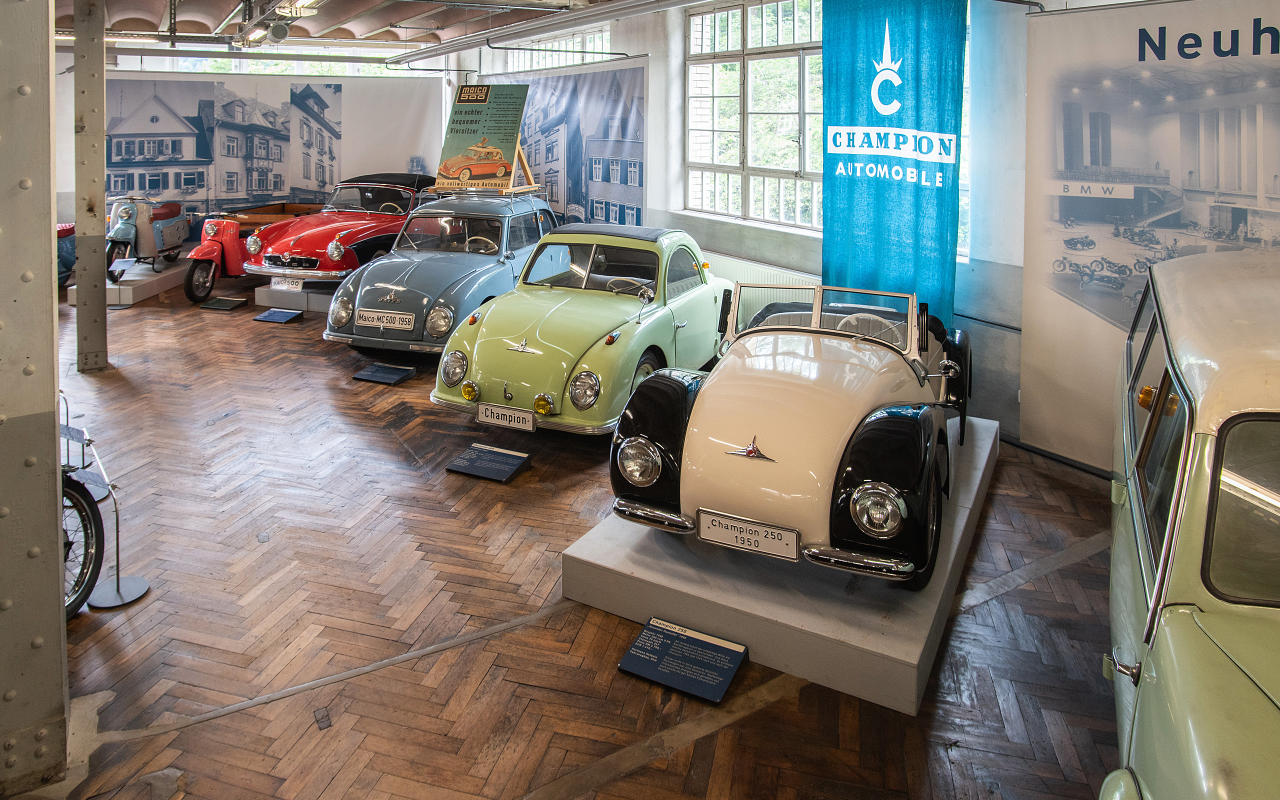 Im Bild sind vier klassische Automobile in einem Ausstellungsraum zu sehen. Die Autos sind in verschiedenen Farben: rot, blau, grün und weiß/ schwarz. Die Modelle sind mit Schildern versehen, die ihre Marken und Baujahre anzeigen. Im Hintergrund sind Fenster und Banner zu erkennen.