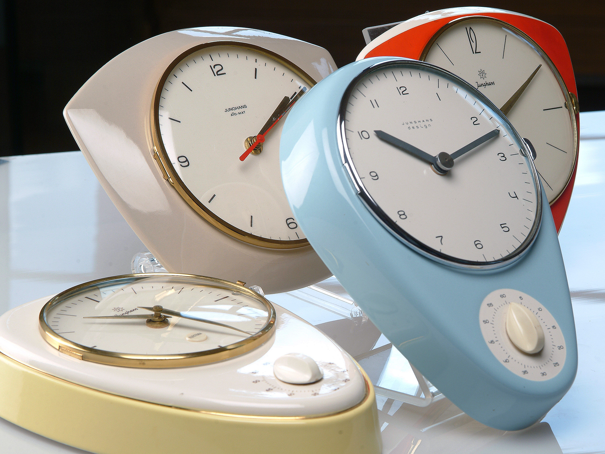 Vier verschiedene Küchenuhren stehen auf einem Tisch. Die Uhren haben unterschiedliche Farben und Designs: eine beige, eine rote, eine blaue und eine weiße Uhr. Die Ziffern sind klar ablesbar, und einige haben einen zusätzlichen Drehknopf. Sie wirken modern und dekorativ.