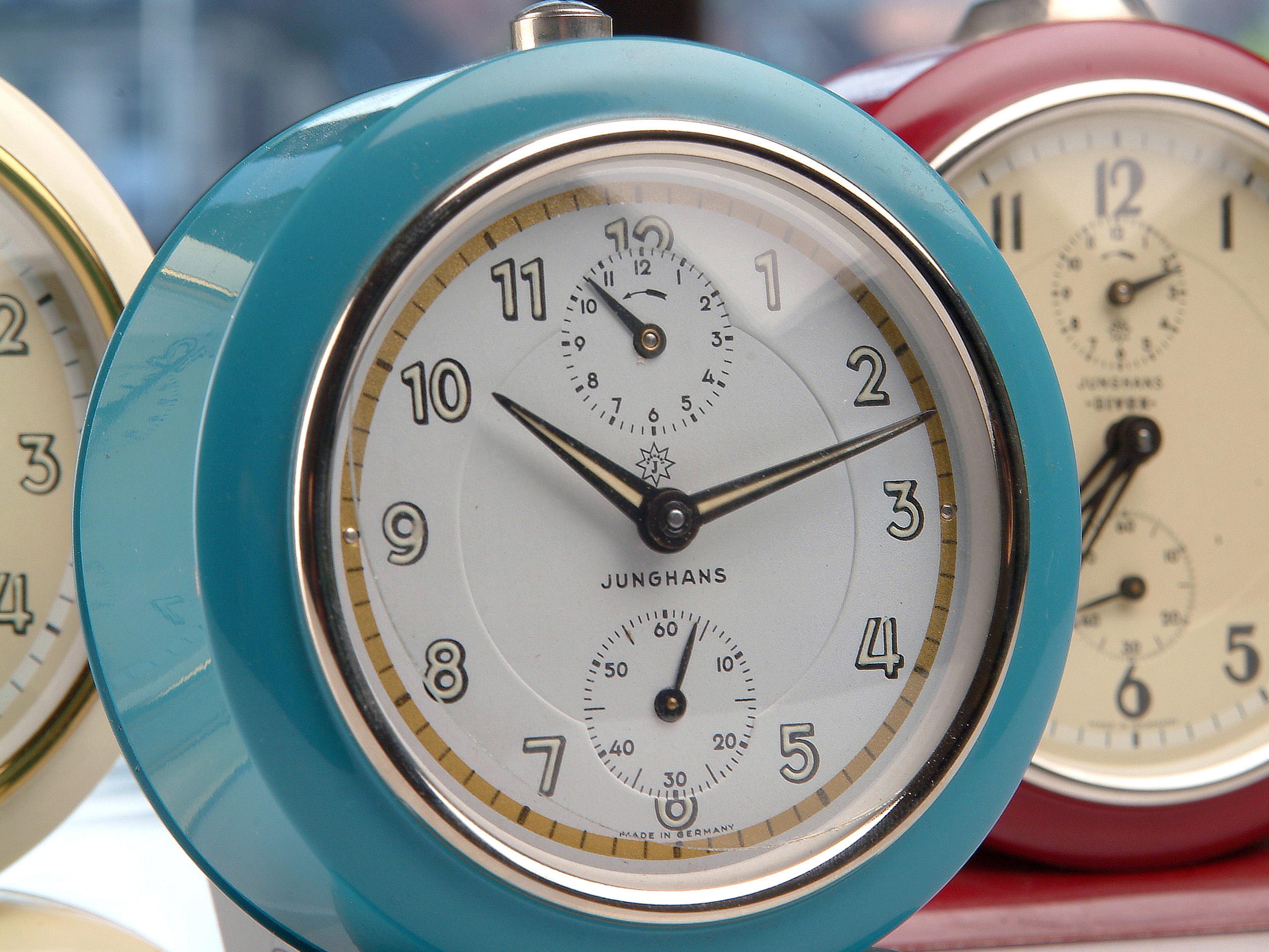 Drei Wecker in verschiedenen Farben stehen nebeneinander. Der vorderste ist in einem leuchtenden Türkis gehalten und hat römische Ziffern auf dem Ziffernblatt. Es gibt zusätzlich einen kleinen Sekundenzeiger. Die Marke Junghans ist sichtbar.