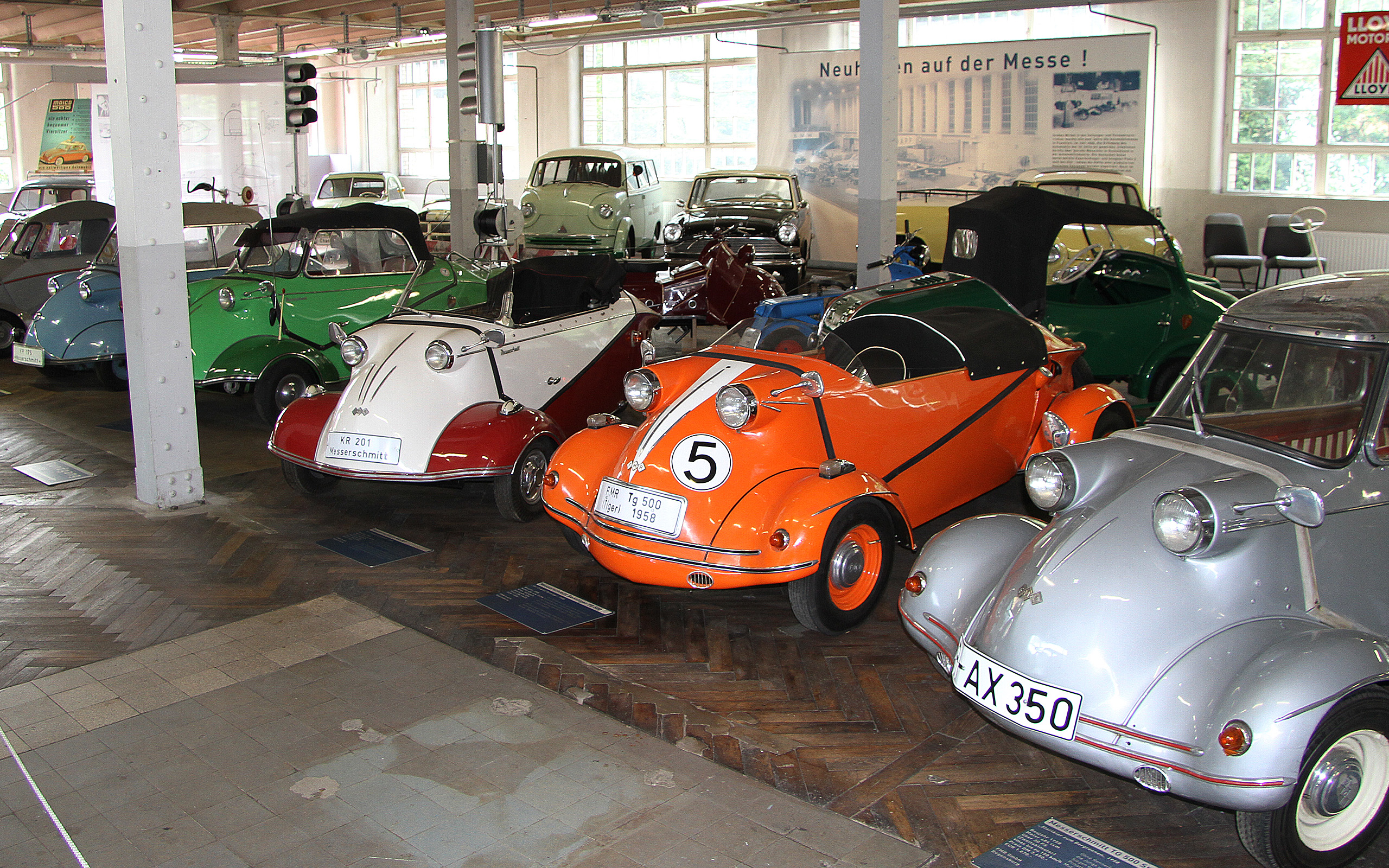 In einem Automuseum stehen mehrere Vintage-Fahrzeuge in einer Reihe. Die Autos sind bunt lackiert, darunter ein auffälliger orangefarbener Kleinwagen mit der Nummer 5. Im Hintergrund sind weitere Oldtimer und eine helle, gut beleuchtete Ausstellung zu sehen.