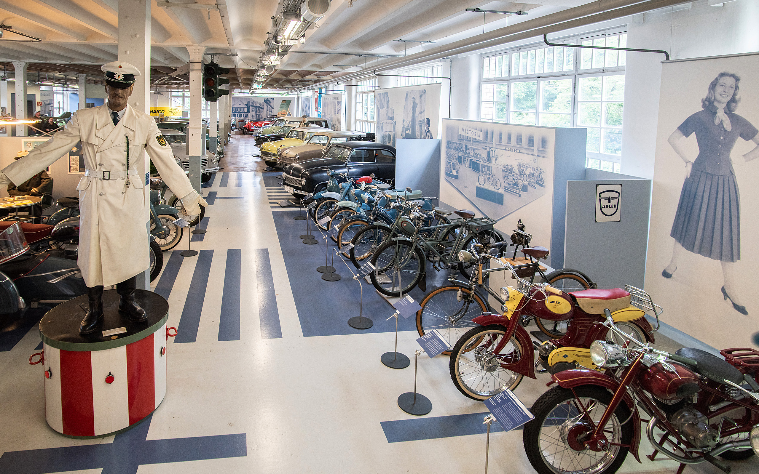 Die Darstellung zeigt einen historischen Ausstellungsraum mit einer Vielzahl von Fahrzeugen, darunter Autos und Fahrräder. In der Mitte steht eine Figur eines Verkehrspolizisten. An den Wänden befinden sich informative Displays über die Geschichte der Mobilität.