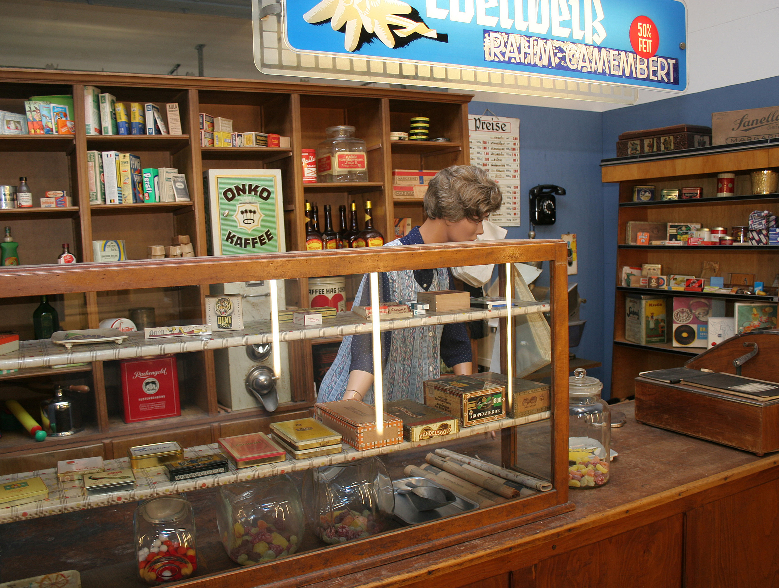 In einem historischen Laden sieht man eine Verkäuferin hinter der Holztheke. Im Regal stehen verschiedene Produkte und Lebensmittel in klassischen Verpackungen. Auf der Theke sind Gläser mit Süßigkeiten und einige kleine Kisten zu sehen. Die Szene vermittelt den Charme eines alten Ladens.