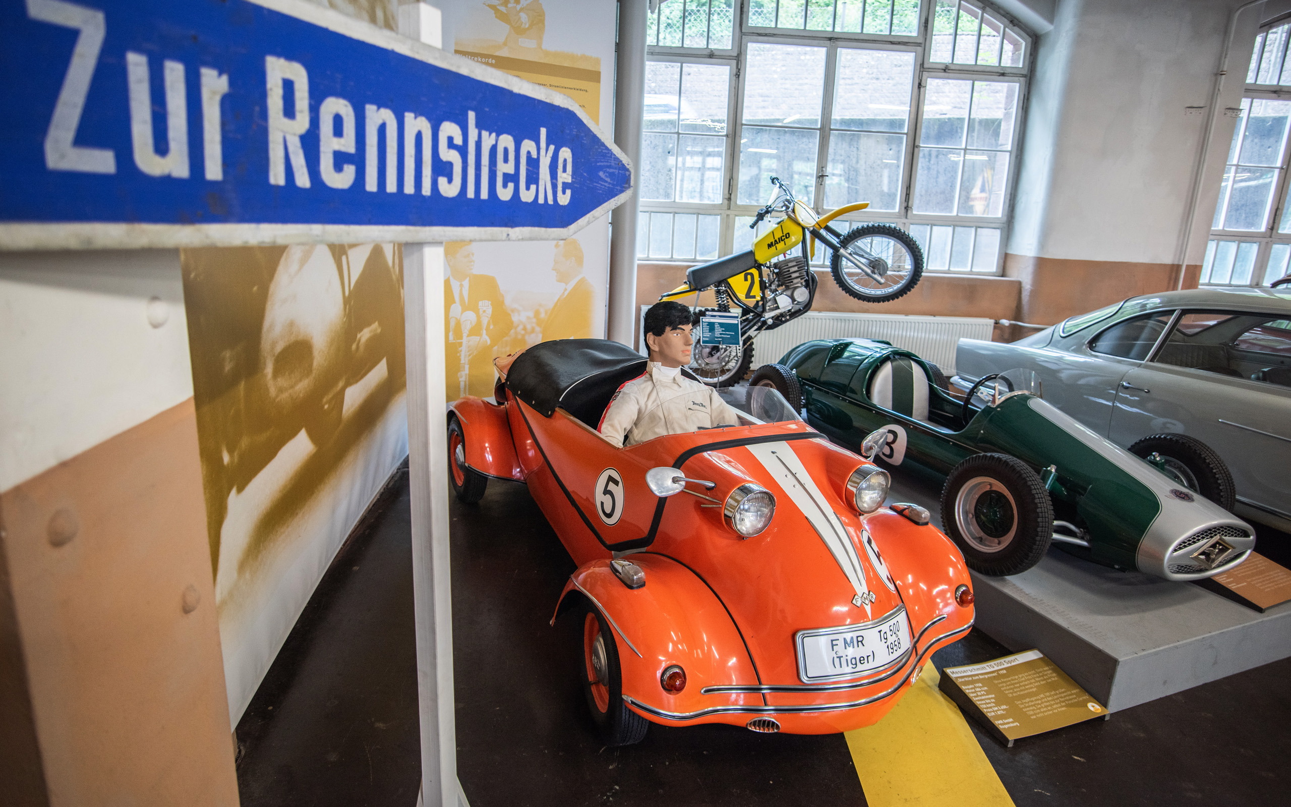 In einem Automuseum steht ein orangefarbenes Dreirad mit einem Fahrer aus Wachs, der einen weißen Rennanzug trägt. Im Hintergrund sind mehrere historische Rennfahrzeuge und Motorräder zu sehen. Ein Schild weist auf die Richtung zur Rennstrecke hin.