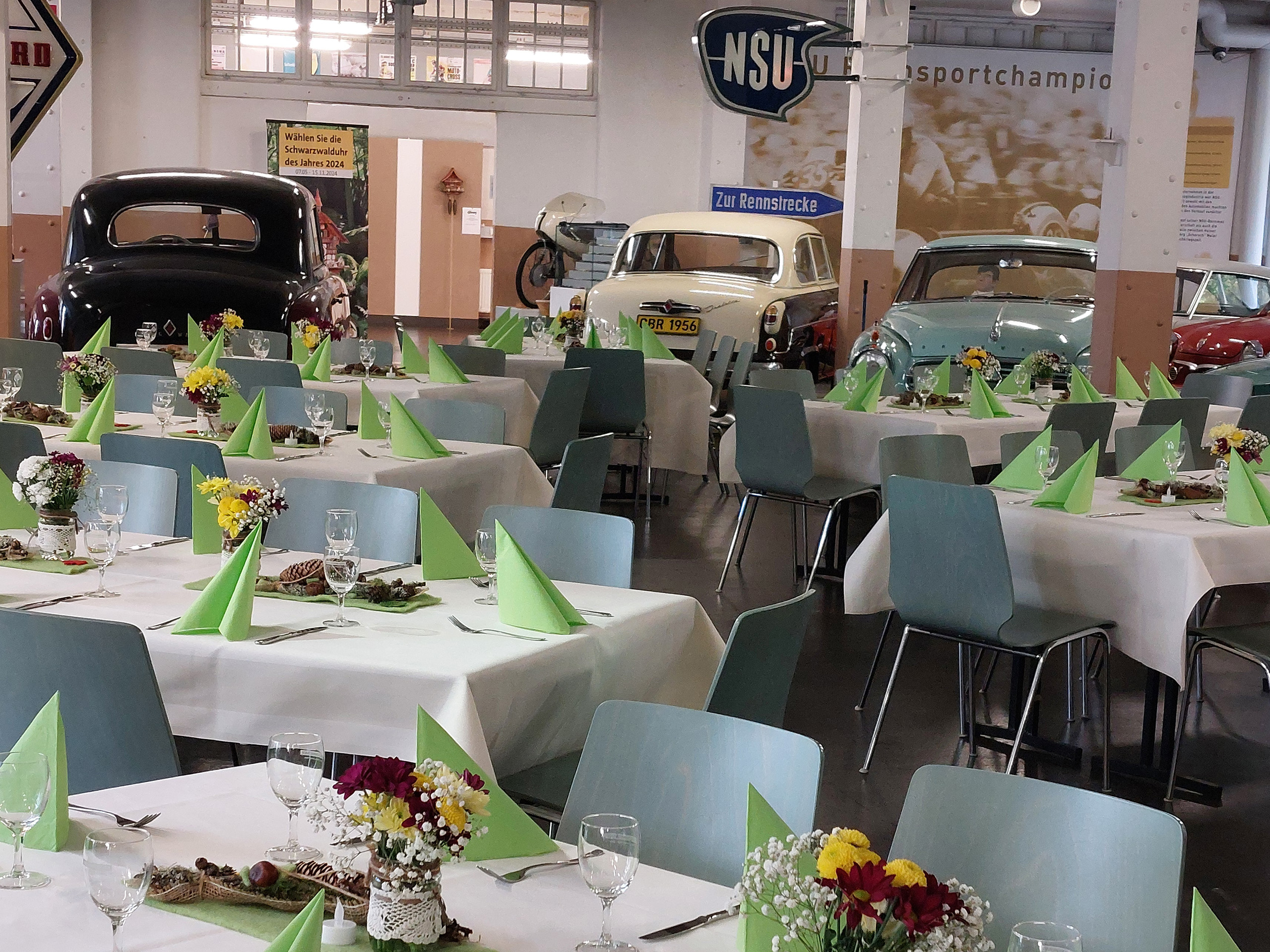Ein festlich gedeckter Saal mit mehreren Tischen. Jeder Tisch hat grüne Servietten, Gläser und kleine Blumengestecke. Im Hintergrund sind einige Oldtimer-Fahrzeuge zu sehen. Die Atmosphäre wirkt einladend und geschmackvoll, ideal für eine Veranstaltung.