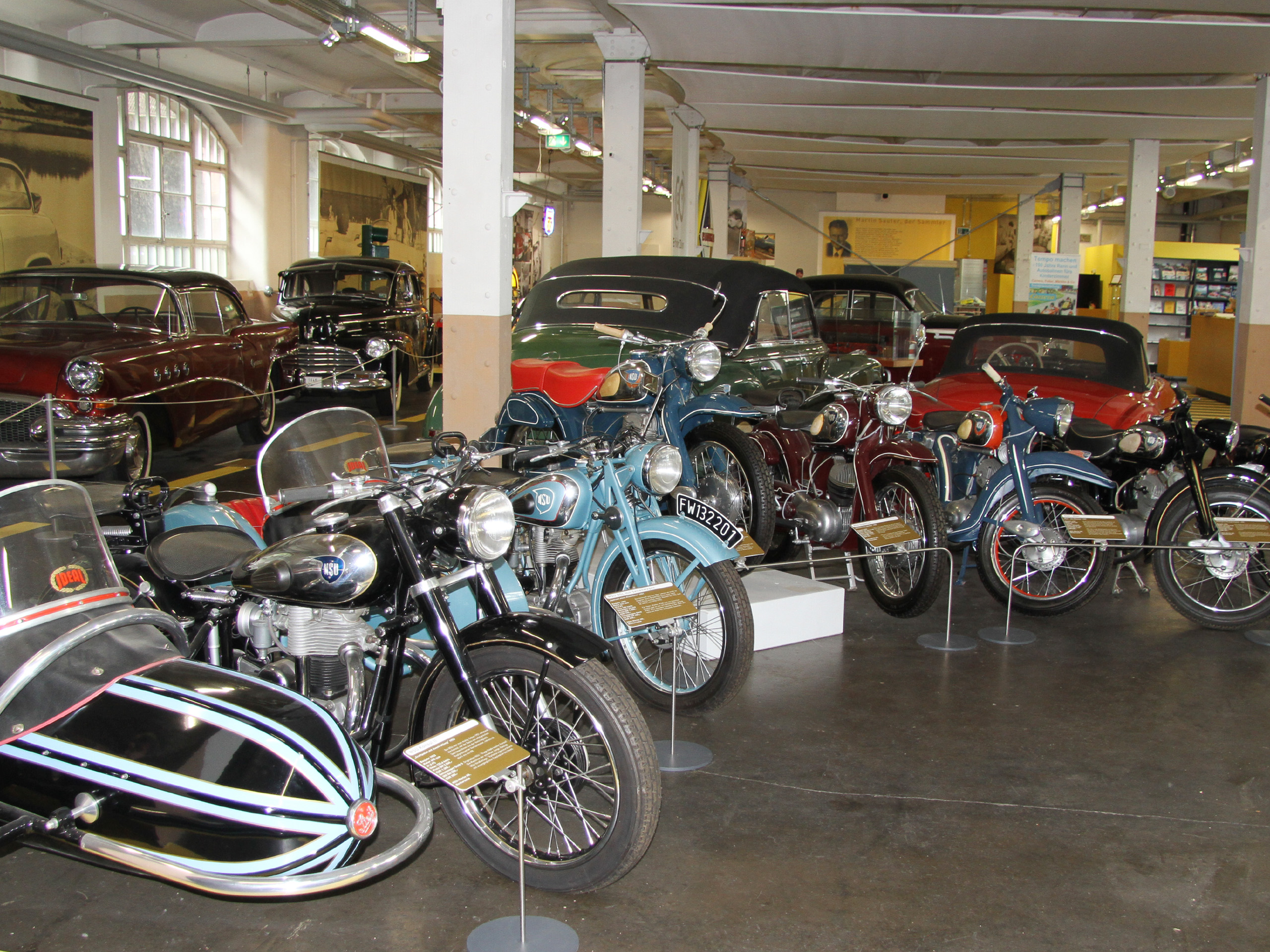 In einem Automuseum sind mehrere historische Motorräder mit Beiwagen ausgestellt. Die Motorräder sind in verschiedenen Farben, darunter Schwarz, Blau und Rot. Im Hintergrund sind einige klassische Autos sichtbar, die die Atmosphäre eines Oldtimermuseums vermitteln.