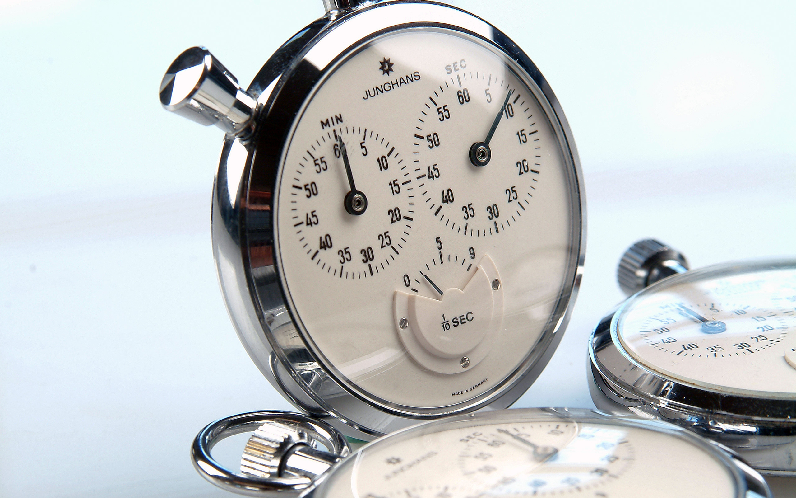 Das Bild zeigt eine Gruppe von Stoppuhren mit silbernem Gehäuse und weißen Zifferblättern. Eine Uhr ist im Vordergrund, die Minuten- und Sekundenzeiger deutlich sichtbar. Die anderen Uhren sind verschwommen im Hintergrund. Alle Uhren sind auf das Zeitmessen ausgelegt.