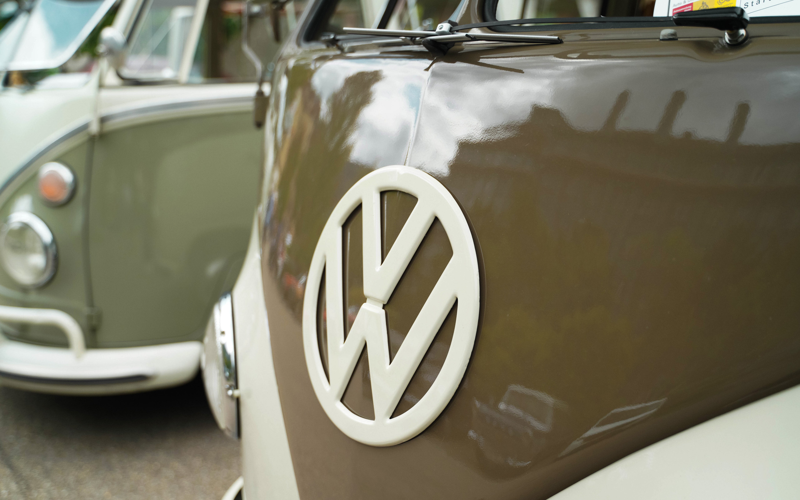 Nahaufnahme des VW-Logos auf einem braunen Fahrzeug. Im Hintergrund sind weitere Fahrzeuge in einer unscharfen Darstellung zu sehen. Die glänzende Oberfläche des Autos reflektiert Licht und Umgebung, während der Fokus auf dem Logo liegt.