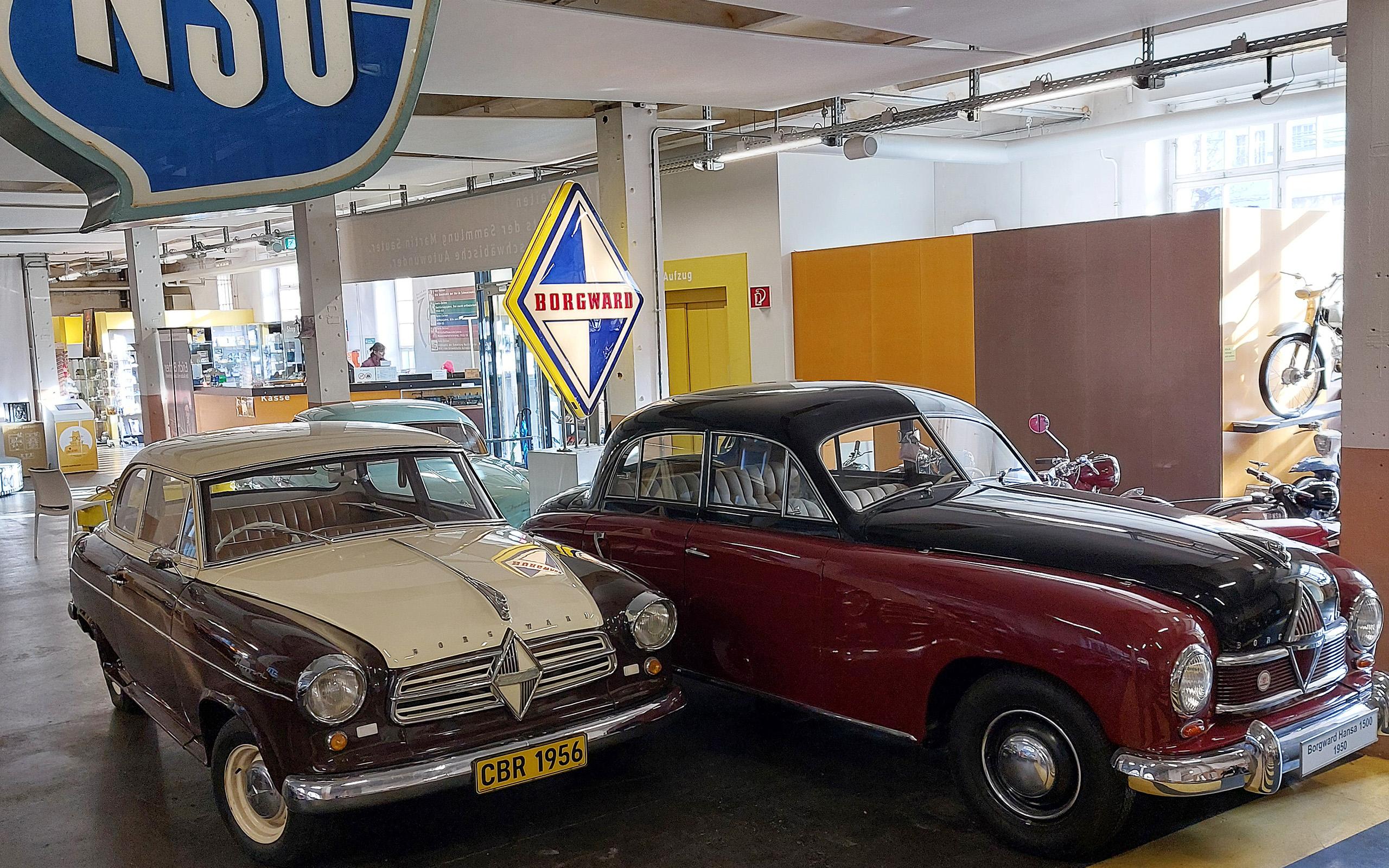 Zwei Oldtimer stehen in einem Museum. Auf der linken Seite ein cremefarbener Wagen mit braunem Dach und dem Kennzeichen „CBR 1956“. Rechts ein roter Wagen mit schwarzem Dach. Im Hintergrund sind Vintage-Schilder von NSU und Borgward sichtbar.