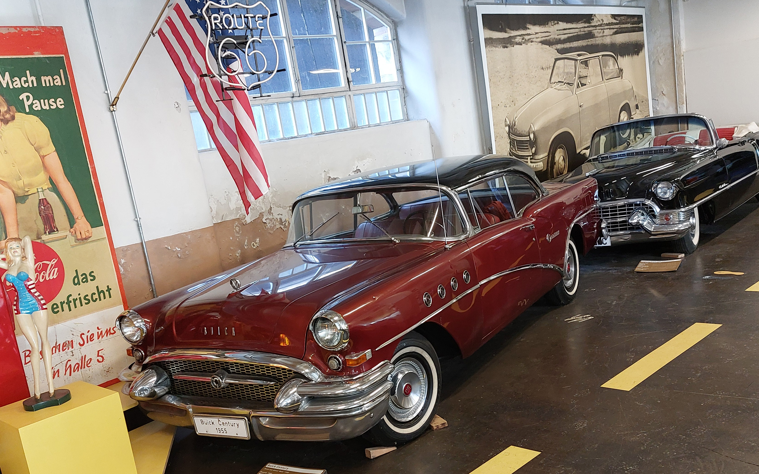 Im Bild sind zwei klassische Autos in einem Museum zu sehen. Das vordere Auto ist ein roter Buick Century von 1955, während das hintere ein schwarzes Cabriolet ist. Im Hintergrund hängen eine amerikanische Flagge und ein Schild mit der Aufschrift: Route 66.
