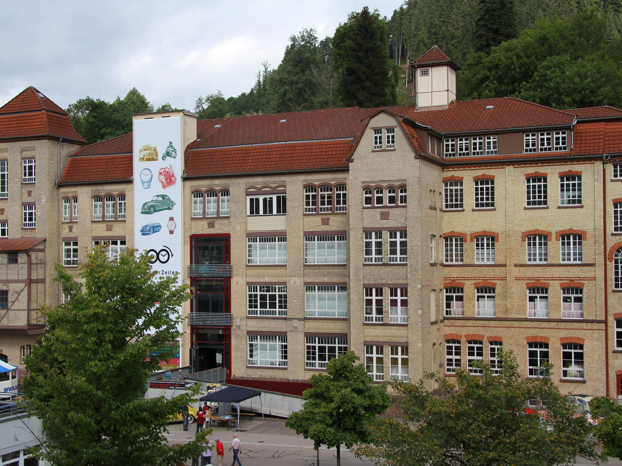 Das Bild zeigt ein historisches Gebäude mit roten Dächern und großen Fenstern. An der Fassade ist ein Werbebanner mit verschiedenen ikonischen Gegenständen, darunter ein Auto und eine Uhr, angebracht. Im Vordergrund sind Bäume und Menschen zu sehen.