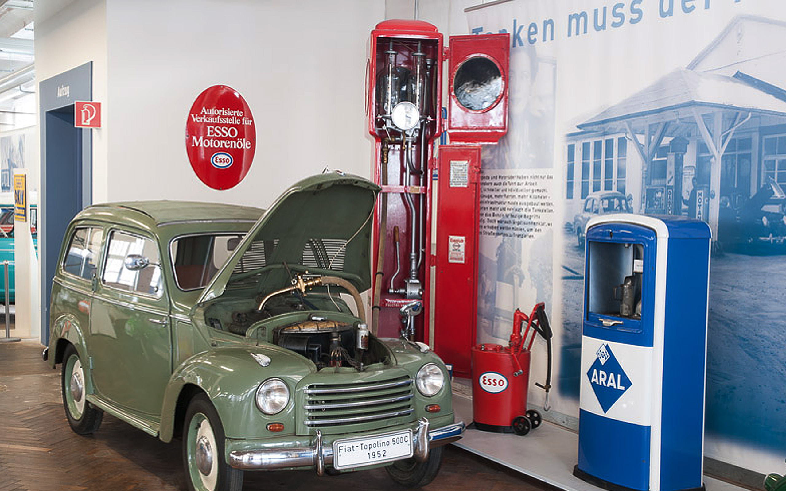 Ein grüner Fiat Topolino von 1952 mit offenem Motorhaube steht ausgestellt. Neben ihm befinden sich alte Zapfsäulen von Esso und Aral, die den historischen Kontext der Automobilkultur verdeutlichen. Der Hintergrund zeigt eine Wand mit textlicher Information.